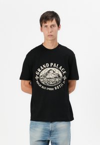 T-shirt nera in cotone con stampa grafica di una montagna e la scritta "GRAND PALACE REPLAY BELLE EPOQUE HOTEL" in crema. Maniche corte.