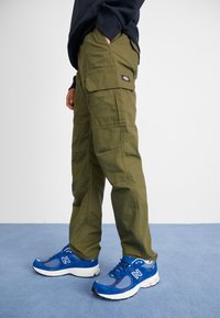 Olivgröna cargobyxor med sidofickor, tillverkade av slitstarkt material, kombinerade med blåa träningssneakers med vita detaljer och en strukturerad sula.