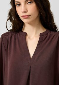 Vrouw met lang donker haar, gouden oorringen en een donkerbruine blouse met een V-hals en gerimpelde schouders.