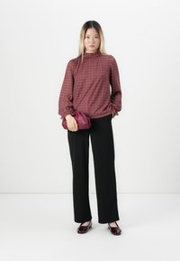 Vrouw staat in een geprinte bordeauxblauwe blouse, zwarte wijde broek, houdt een bijpassende bordeauxblauwe clutch vast en draagt bordeaux laklederen schoenen.