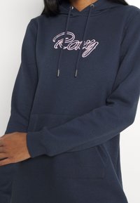Sudadera azul marino hecha de un tejido de textura suave, con un logo "Roxy" bordado en rosa, capucha ajustable con cordón y bolsillo frontal.