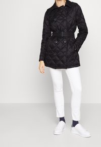 Svart vadderad kappa med knytmidja, dubbelknäppta knappar och krage. Kombinerad med vita byxor och vita sneakers.