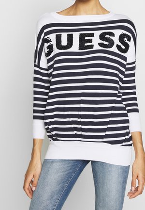 Woman wearing a black and white striped sweater with "GUESS" sequined across the chest and light blue jeans.