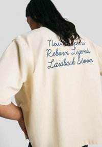 Persona con lunghi capelli scuri che indossa una camicia crema con testo ricamato blu: "Nuovi Capitoli Rinascenti Leggende Storie Rilassate."