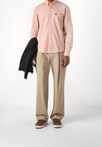 Helle rosa Langarm-Hemd mit Knopfleiste und TaschenDetail, kombiniert mit lockeren beigen Hosen und braunen Turnschuhen. Schwarze Jacke über einem Arm drapiert.