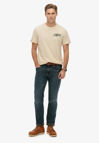 Camiseta de algodón beige con gráficos impresos, jeans de mezclilla azul y botas de cuero marrón. El modelo posa con las manos en los bolsillos.