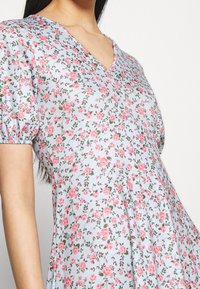 Blommig V-ringad klänning i ljusblått tyg, prydd med rosa blommor och gröna blad. Kort puffärmar och en utsvängd kjoldesign.