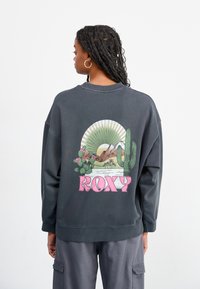 Mørk grå hettegenser med avslappet passform, med et fargerikt ørkenlandskap grafikk og ordet "ROXY" i rosa bak.