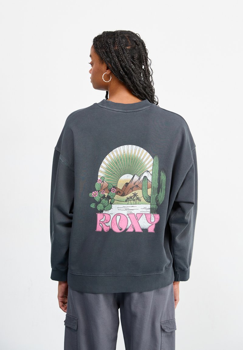 Mørk grå hettegenser med avslappet passform, med et fargerikt ørkenlandskap grafikk og ordet "ROXY" i rosa bak.