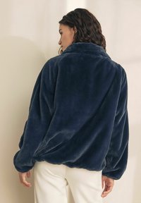 Jachetă din fleece supradimensionată de culoare bleumarin, cu guler, umeri căzuți și textură moale. Are mâneci largi și o siluetă relaxată.