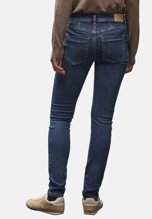 Jeans skinny en denim bleu foncé, texture lisse, taille mi-haute, poches arrière classiques, associés à des baskets beige clair.