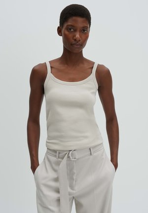 Femme aux cheveux courts portant un débardeur côtelé beige et un pantalon beige taille haute avec une ceinture en tissu, debout les mains dans les poches.