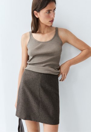 Kvinde iført en ribbet taupe tanktop og en brun tekstureret nederdel, står mod en hvid væg med den ene hånd på hoften.