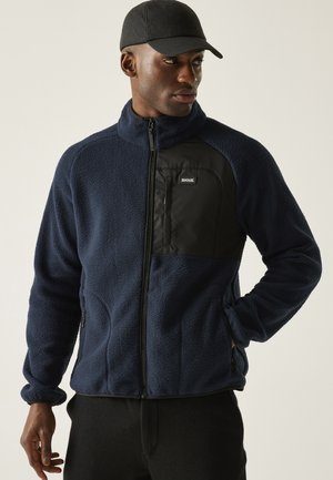 Man draagt een marineblauwe fleecejas met een zwart borstzakje en een zwarte rits, gecombineerd met een zwarte pet en zwarte broek, en kijkt naar rechts.