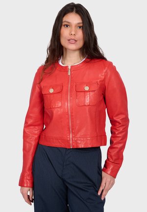Žena stojící před jednobarevným pozadím, oblečená v červené kožené bundě na zip s dvěma knoflíkovými kapsami na prsou a tmavých kalhotách s proužky.