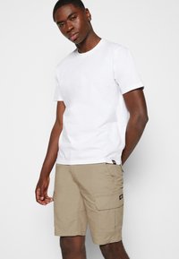 T-shirt en coton blanc léger associé à un short cargo beige avec plusieurs poches et une texture subtile. Présentation d'une tenue décontractée.