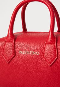 Punane nahast käekott, millel on tekstureeritud pind, ümarad kahekordsed käepidemed ja kuldne "VALENTINO" logo esiküljel. Kompaktne kuju, millel on struktureeritud disain.