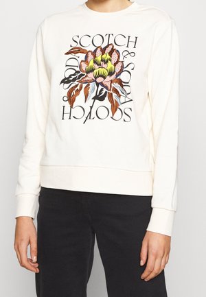 Osoba ubrana w biały sweter z dużym, haftowanym kwiatem o brązowych, żółtych i różowych płatkach oraz czarnym napisem "Scotch & Soda".