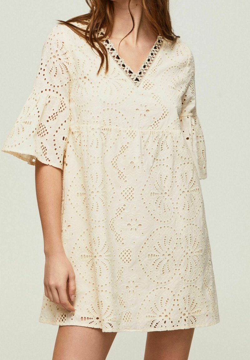 Femme portant une robe courte de couleur crème avec des motifs en dentelle œillet floraux et un décolleté en V avec un bord noir et crème.