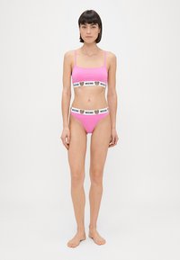 Roza braletka in visoki bikini z belimi elastičnimi trakovi, na katerih je logotip medveda. Enostavna, gladka tekstura, minimalističen dizajn.