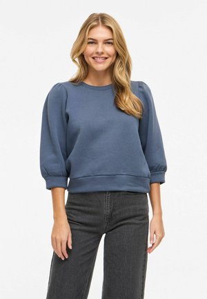 Ung kvinde med langt blondt hår iført en blå sweatshirt med pufærmer og sorte jeans, smilende med hænderne afslappet ved siden.