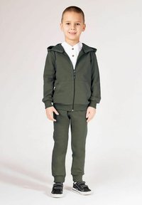 Zielona bluza z kapturem i dopasowane spodnie wykonane z miękkiego materiału. Bluza posiada przedni zamek błyskawiczny i dwie boczne kieszenie. Czarne trampki z białymi podeszwami.