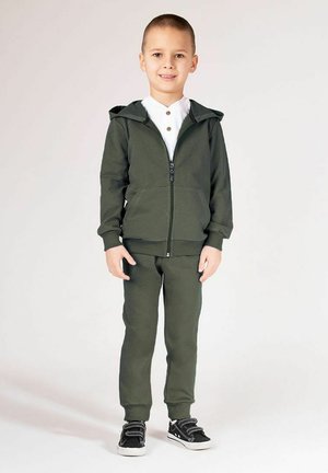 Zielona bluza z kapturem i dopasowane spodnie wykonane z miękkiego materiału. Bluza posiada przedni zamek błyskawiczny i dwie boczne kieszenie. Czarne trampki z białymi podeszwami.