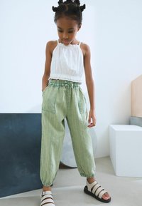 Top blanco sin mangas con dobladillo con volantes, combinado con pantalones ligeros verdes con cordón y rayas verticales, que presentan bolsillos, y usados con sandalias blancas.