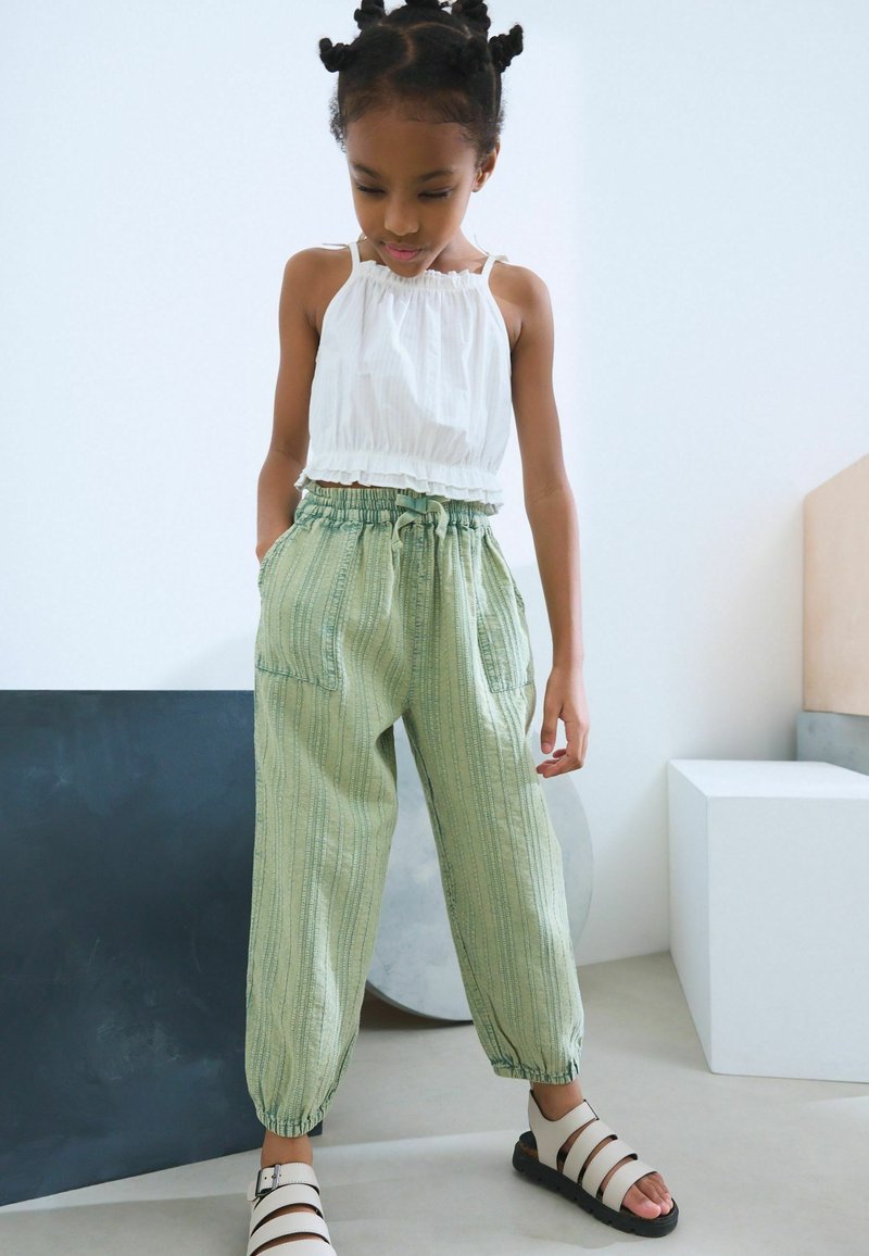 Top blanco sin mangas con dobladillo con volantes, combinado con pantalones ligeros verdes con cordón y rayas verticales, que presentan bolsillos, y usados con sandalias blancas.