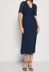 Mörkblå midi-klänning med V-ringning, korta ärmar och figurnära midja. Lätt, flödande tyg med veckad kjol. Svarta remsandaler med klack.