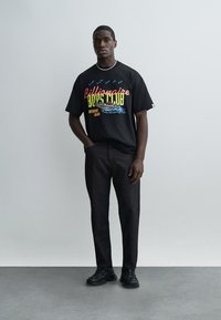 Ung mann som står med den ene hånden i lommen, iført svart T-skjorte med grafisk trykk, mørke bukser, svarte joggesko og et hvitt perlesmykke.