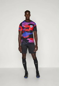 Muž stojí v anglickom futbalovom dresu s viacfarebným červeným, fialovým a čiernym tričkom, čiernymi šortkami, čiernymi ponožkami s bielymi logami Nike a kopačkami.