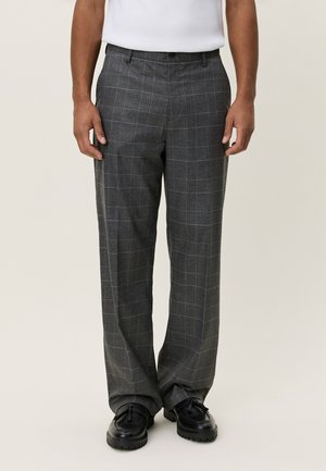 Man die grijze geruite broek, zwarte loafers met kwastjes en een ingestopt wit overhemd draagt, staand tegen een effen lichte achtergrond.