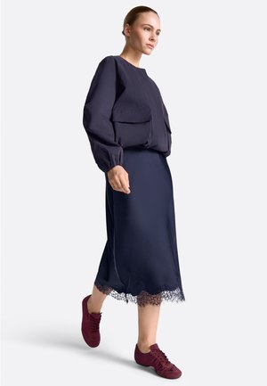 Nő sétál, sötétkék hosszú ujjú dzsekit, sötétkék midi szoknyát csipkés szegéllyel és bordó fűzős cipőt visel, egyszínű háttér előtt.