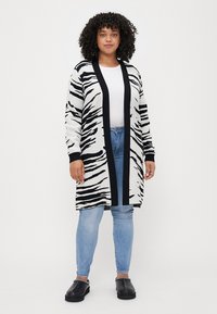 Zebra-striped long cardigan in black and white with a solid black border. Worn over a white top and blue jeans. Made from a soft knit material.
