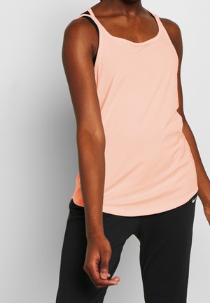 Femme portant un haut sans manches couleur pêche clair par-dessus un soutien-gorge de sport noir et un pantalon Nike noir, posant devant un fond blanc.