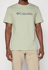 Svetle zelené bavlněné tričko s logem "Columbia" v tmavě šedé barvě, kulatý výstřih a krátké rukávy. Kombinováno se světle béžovými cargo šortkami.