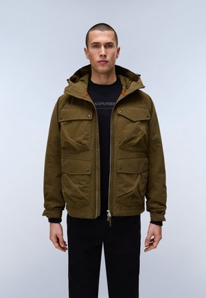 Mand iført olivengrøn hættetrøje over sort skjorte og sorte bukser, stående mod en ensartet lys baggrund.