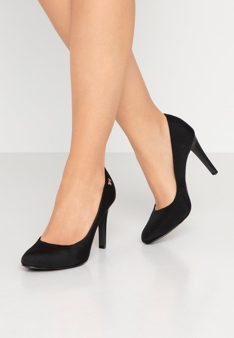 Femme portant des escarpins en daim noir à talons hauts, debout sur un sol clair et brillant, jambes croisées, sur un fond uni clair.
