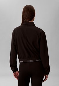 Persona ar gariem brūniem matiem demonstrē melnu jaku un melnas bikses, jakas apakšējā malā ir elastīga lenta ar "Calvin Klein" zīmolu.