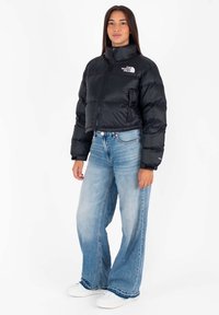 Donna che indossa un giubbotto nero corto in piuma, jeans azzurri a gamba larga e sneakers bianche, in piedi contro uno sfondo bianco uniforme.