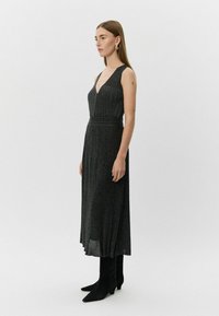 Zwarte vloerlange jurk van textuur met geplooide stof, V-hals en brede banden. Gecombineerd met zwarte, puntige laarzen.