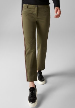 Persona con pantalones de pernera recta color verde oliva con franjas rojas laterales y zapatillas negras con plataforma caminando sobre un fondo gris.