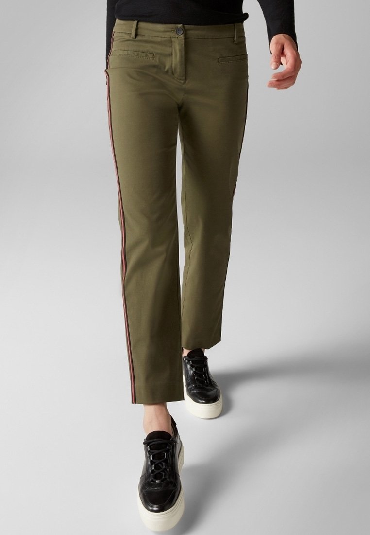 Personne portant un pantalon olive droit avec des bandes latérales rouges et des baskets plateformes noires, marchant sur un fond gris.