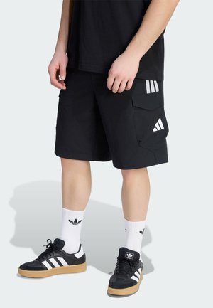 Person iført sorte Adidas-shorts, hvide Adidas-strømper og sorte Adidas-sneakers med hvide striber, der står på en lys baggrund.