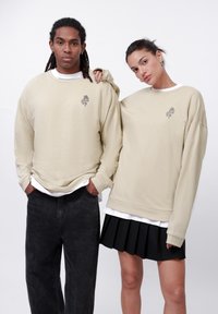 Beige oversized sweatshirts with embroidered design, worn over white layers; paired with black pants and pleated skirt; casual, relaxed fit.