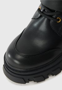 Black leather ankle boot with a textured surface, rounded toe, gold eyelets, and a thick, rugged sole. Features black laces and stitching.