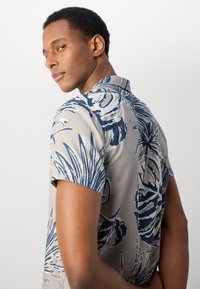 Cămașă cu mânecă scurtă în bej deschis, cu un model de frunze tropicale în nuanțe de bleumarin, având un guler ascuțit și un design drept pentru spate.
