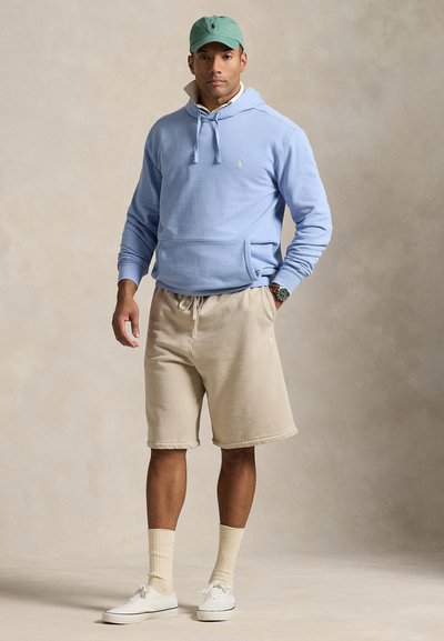 Lyseblå hettegenser med kengurulomme, beige shorts, hvite joggesko og grønn caps med logo. Avslappet, mykt stoff og ledig passform.