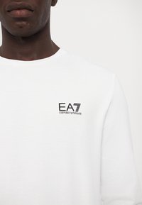 Hvit langermet t-skjorte laget av bomull, med en svart EA7 Emporio Armani-logo på venstre brystområde. Glatt tekstur, rund hals.